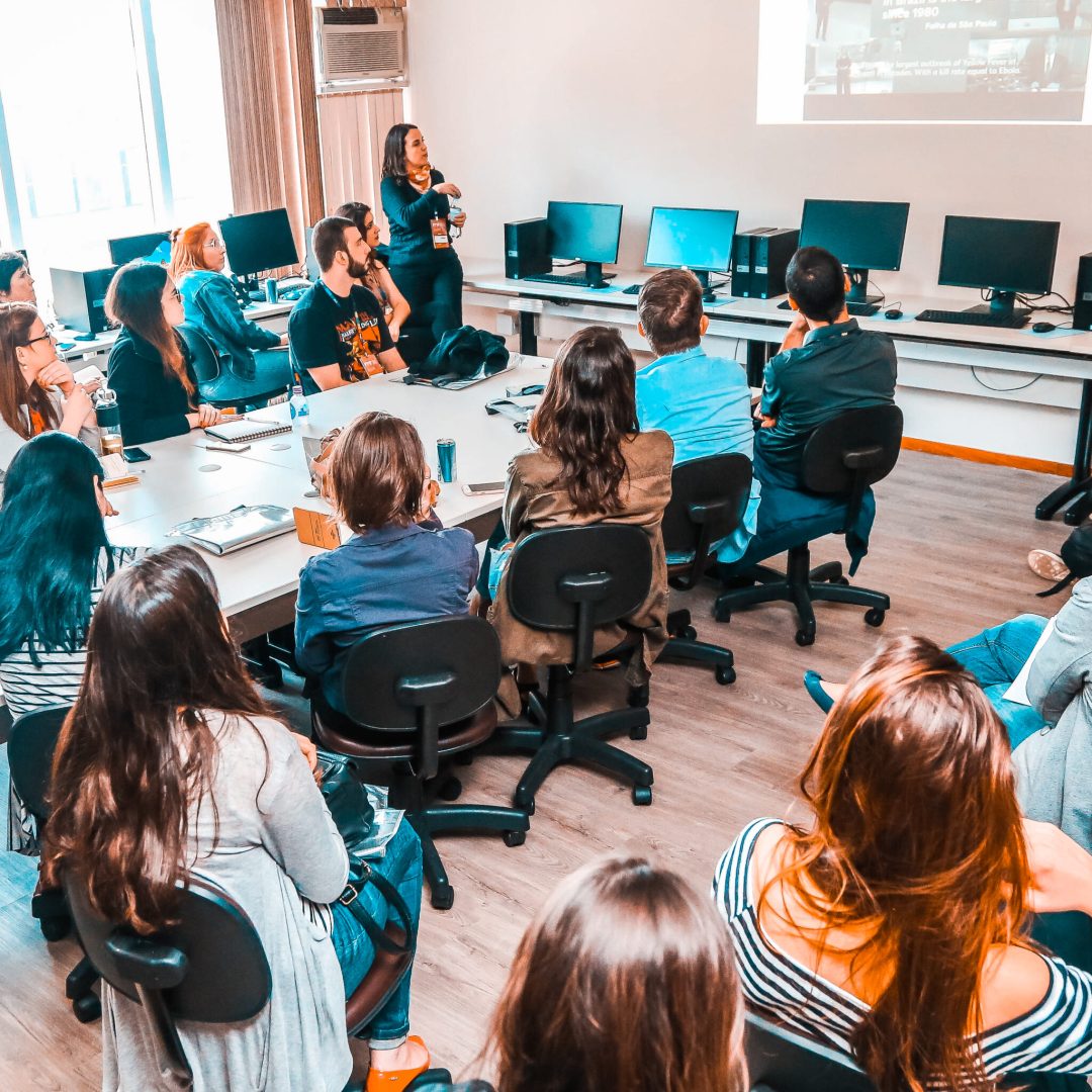 #PraCegoVer #PraTodesVerem: A foto mostra diversas pessoas sentadas em cadeiras assistindo a uma palestra, que está sendo apresentada pela criadora da Otimifica, Ariane Feijó. Ela está de pé, ao lado de uma projeção que está sendo feita na parede.